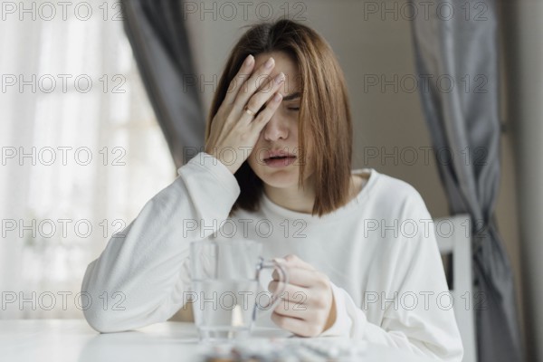 Woman holding her forehead, looking unwell, with a glass of water and curtains in the background