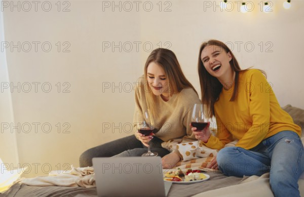 Two cute happy funny women friends having fun together, arranging home cinema evening, watching comedy on laptop, laughing out loudly, sitting on bed with cheese plate and glasses of red wine