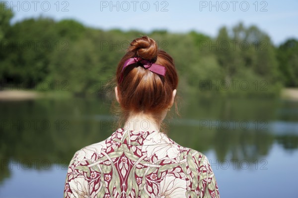 back view of unrecognizable young woman with floral dress and red hair bun looking at lake in solitude