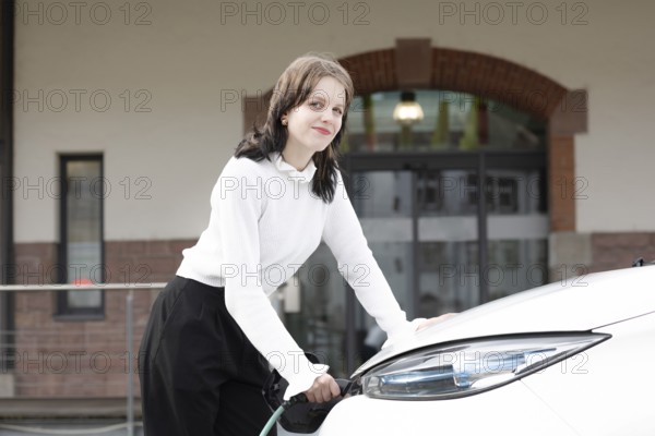 Friendly woman in white jumper and smile leaning on a charging electric car with urban background