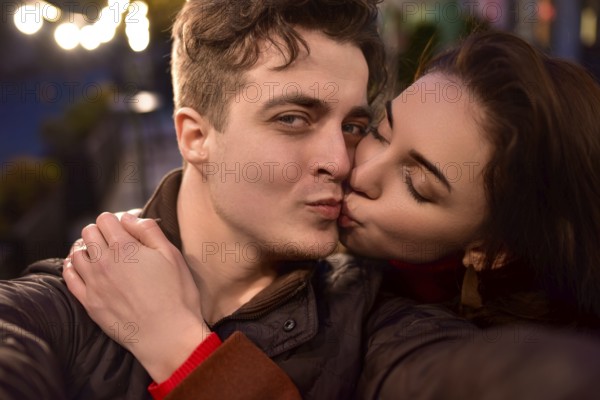A young trendy couple in love walks down a night street and takes selfies. The girl is kissing her boyfriend