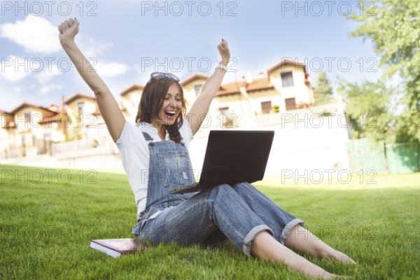 Female gets joyful emotions by talking to her friends on a conference call from notebook, she dressed in jeans overalls sitting on grass in the park