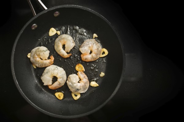 Fresh shrimps are sauteed with garlic in olive oil in a cooking pan on the black stove, preparation for a delicious appetizer, copy space, high angle view from above, selected focus