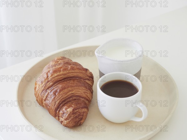 Simple breakfast on a tray on the white table. Croissant, coffee cup and milk jug