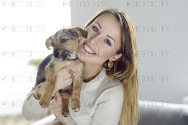 Pretty happy young woman with her pet dog held up to her cheek in her hands in a natural tender portrait indoors on a sofa