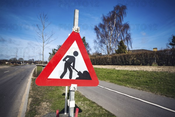 Road construction sign by a roadside in a city on a sunny day with blue sky