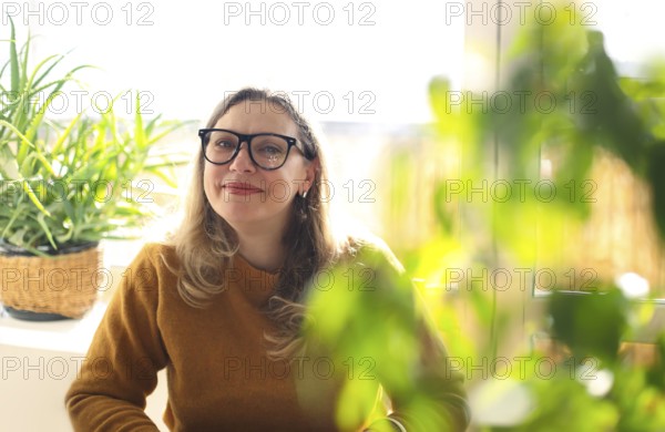 Portrait of smiling middle-aged woman wearing glasses sitting in room with green potted houseplants, selective focus. Positive happy 40s female teacher in eyeglasses in greenery, working in school
