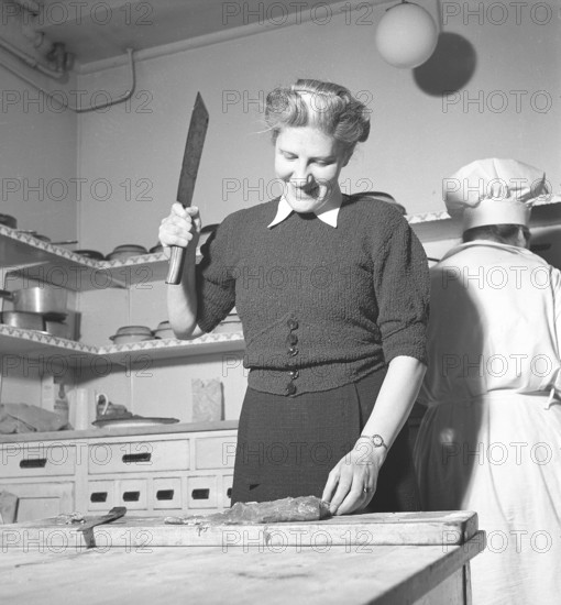 Happy woman in the kitchen in 1948. A woman in a restaurant kitchen stands and cuts the meat into pieces with a larger knife. She has placed the piece of meat on a cutting board and has raised the butcher knife to begin. Sweden 1948. Kristoffersson ref AF26-4