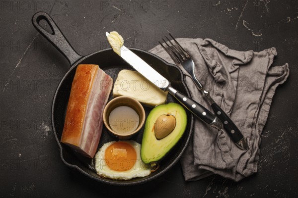 Main keto foods as butter, olive oil, fried egg, avocado, fat meat bacon for ketogenic diet on black cast iron pan on dark rustic stone background, products for healthy weight loss top view