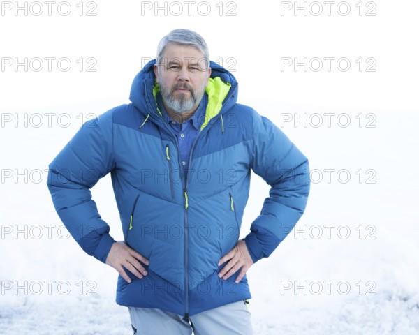 Man, 56 years old with grey hair and a full grey beard standing in front of a snowy area in winter, facing the camera, hands on hips, critical look, thick down anorak, down jacket, blue shirt, white background