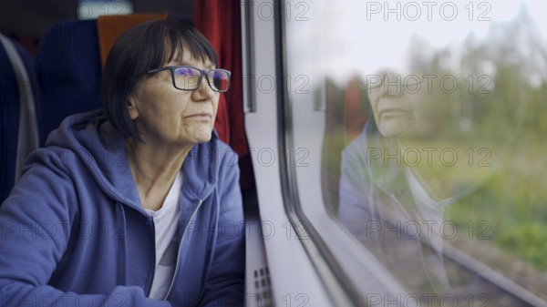Elderly lady in glasses travels in train and looking out the window reflected in the glass