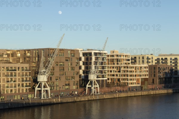 Weather, winter, moon, walkers and historic harbour cranes in front of modern architecture in the sunny evening light on the Elbe on the Versmannkai promenade, Baakenhafen district, Hafencity, Hamburg, Germany, Europe