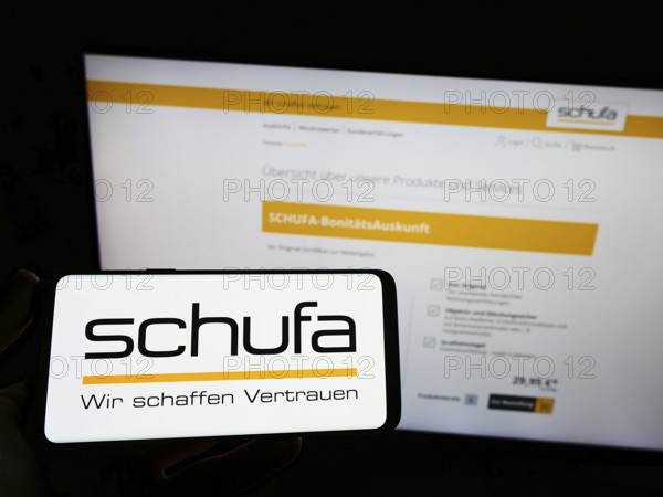 Stuttgart, Germany, 02-13-2022: Person holding mobile phone with logo of German credit protection company Schufa Holding AG on screen in front of webpage. Focus on phone display, Europe