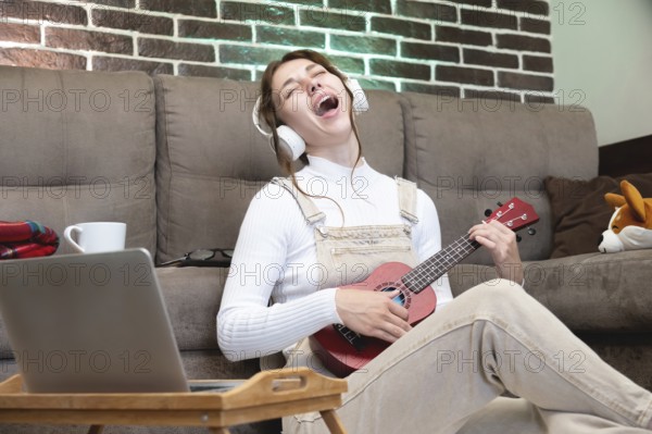 Remote music lessons, playing the ukulele or teaching vocals, singing. Hipster student recording lesson or studying using laptop webcam while sitting on floor in living room