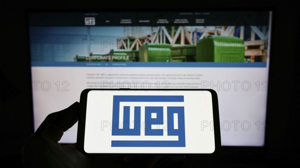 Stuttgart, Germany, 07-23-2022: Person holding mobile phone with logo of Brazilian company WEG Equipamentos Eletricos S.A. on screen in front of web page. Focus on phone display, Europe