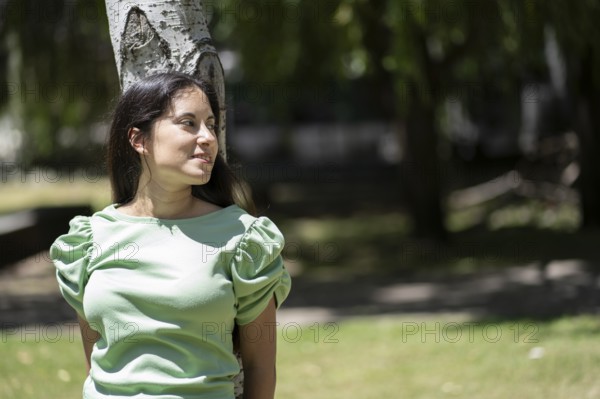 Latina woman in a park leaning against a tree looking to the side