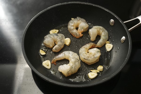 Raw peeled shrimps and garlic are sauteed in a cooking pan on the black stove, preparation for a delicious appetizer, copy space, selected focus, narrow depth of field
