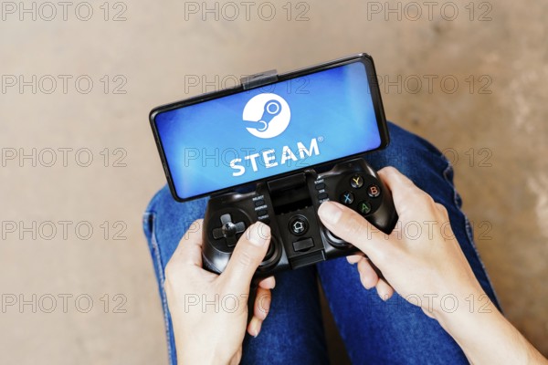 In this photo illustration, a person playing on a joystick and the Steam logo displayed on a smartphone screen