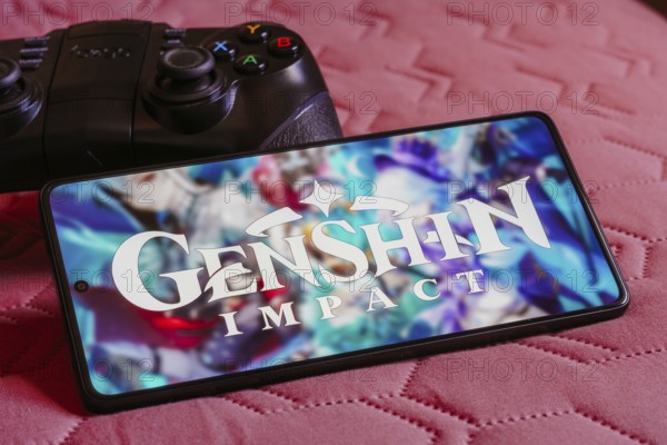 In this photo illustration, the Genshin Impact logo is displayed on a smartphone screen, next to a gamepad