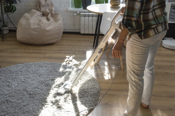 A woman vacuums a round carpet in a house among house plants with a hand vacuum cleaner. General cleaning of the house, cleaning service and housewife