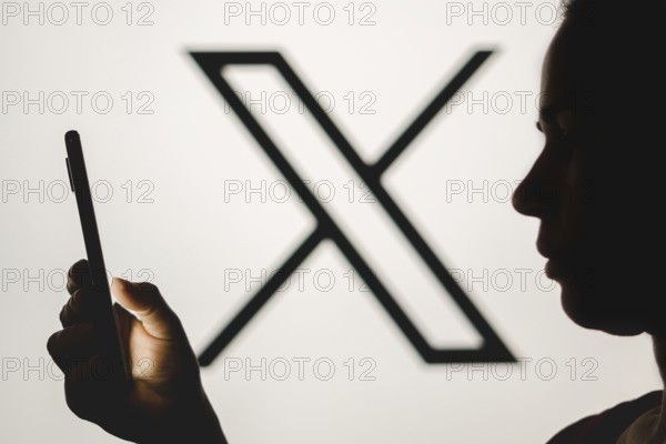 In this photo illustration, a woman's silhouette holds a smartphone with the Twitter (X) new logo in the background
