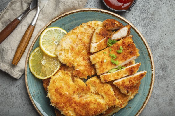 Crispy panko breaded fried chicken fillet with green salad and lemon cut on plate on gray rustic concrete background table with ketchup from above. Japanese style deep fried coated chicken breasts, food photography, food photography