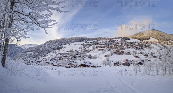 Winter panorama of snow covered La Clusaz village nestled in a hilly landscape in Annecy, France. Wooden cabins are scattered across the snowy hillside, surrounded by snow laden trees