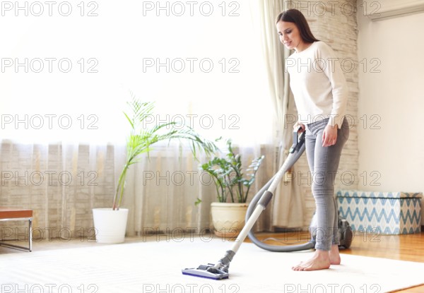 Full body young female using vacuum cleaner to tidy carpet in light room at home