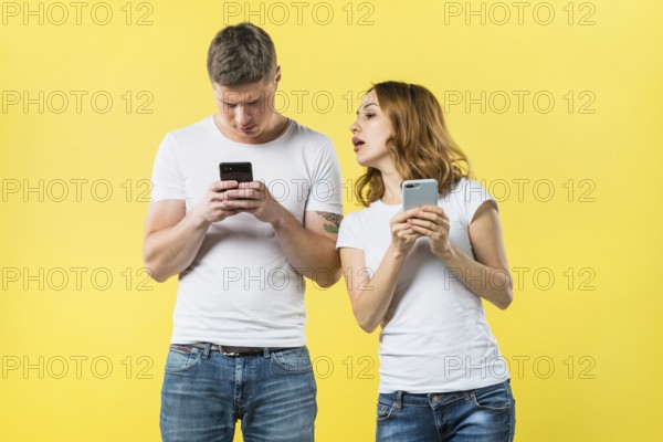 Suspicious girlfriend spying his boyfriend texting mobile phone against yellow backdrop