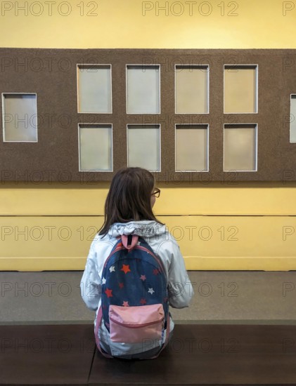 Back view of teen girl with backpack in museum visiting exhibition, schoolgirl sitting on bench in art gallery and looking with interest at art objects or exhibits on wall