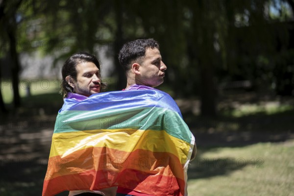 Couple of gay latin men walking in a park wrapped in the LGBT flag
