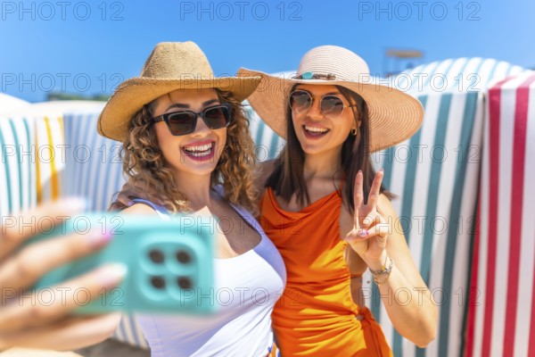 Two cheerful women wearing stylish sunglasses and trendy sun hats are capturing a joyful selfie on a sunny beach, surrounded by sand and sea