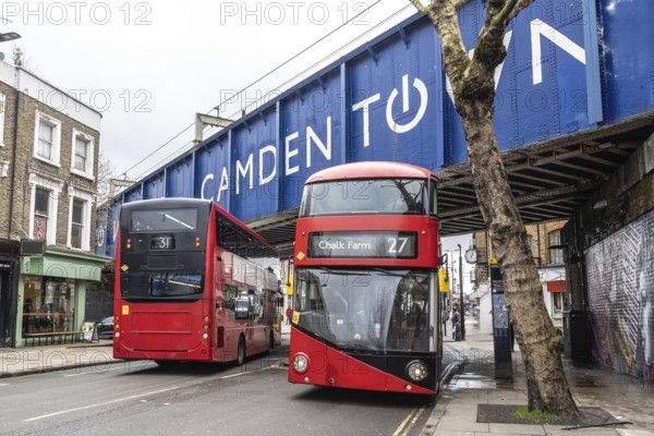 Red double decker buses driving on a london street, passing under a bridge with the camden town sign