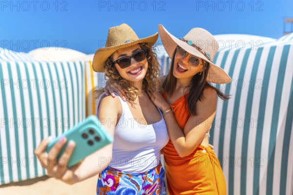 Two cheerful women wearing summer hats and sunglasses are taking a selfie on a sunny beach with striped beach huts in the background