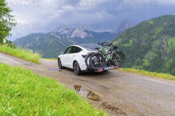 White car with bicycles on road train, surrounded by green mountains and cloudy sky, electric car on holiday, Dolomites, South Tyrol, Dolomites, Italy, Europe