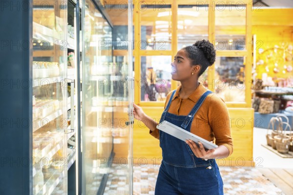 Young supermarket employee opening refrigerated section door and checking food products using digital tablet