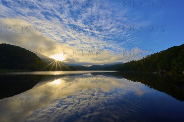 The early morning shows sunbeams through clouds reflecting on a calm lake in a mountainous landscape, summer, Rursee, Obersee, Rurberg, Simmerath, Eifel National Park, Eifel, North Rhine-Westphalia, Germany, Europe