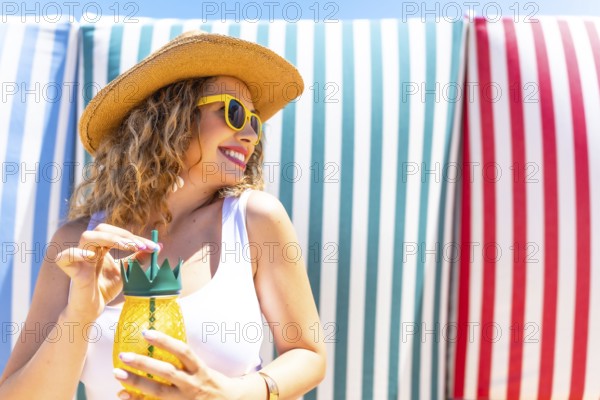 Young woman with straw hat and sunglasses enjoying a refreshing pineapple cocktail on a beach with colorful striped towels in the background, during summer vacation