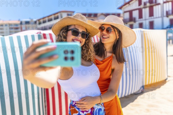 Two smiling women with sun hats and sunglasses taking a selfie on a sunny beach with colorful striped beach huts