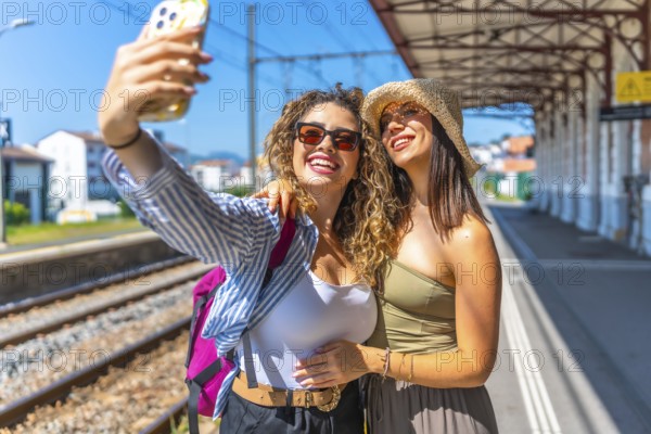 Two happy female tourists taking a selfie with smartphone at a train station, enjoying their summer vacation