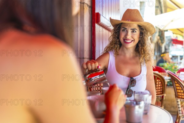 Smiling tourist wearing straw hat pouring milk into coffee, having breakfast with friend at outdoor cafe