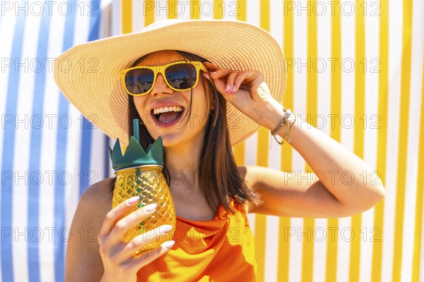 Young woman with sunglasses and a straw hat, enjoying a tropical pineapple juice on a colorful striped background