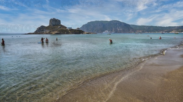 Clear sea with people bathing, surrounded by an island and mountains under a cloudy sky, Agios Stefanos Beach, Agios Stefanos Basilica, Chapel of Agios Nikolaos, Kastri Island, Kos, Dodecanese, Greek Islands, Greece, Europe
