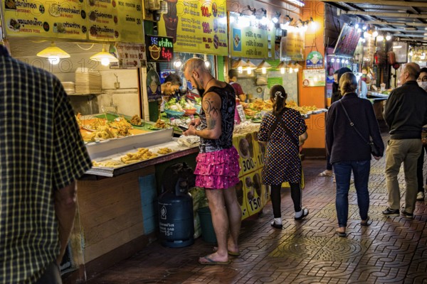 Tatooed man in pink skirt and flip flops ordering food from a street vendor in the Night Market in downtown Chiang Rai, Thailand, Asia