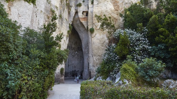 Cave Ear of Dionysus, Overgrown cave entrance with lush vegetation and rocks, people in the distance, Neapolis Archaeological Park, Syracuse, Sicily, Italy, Europe