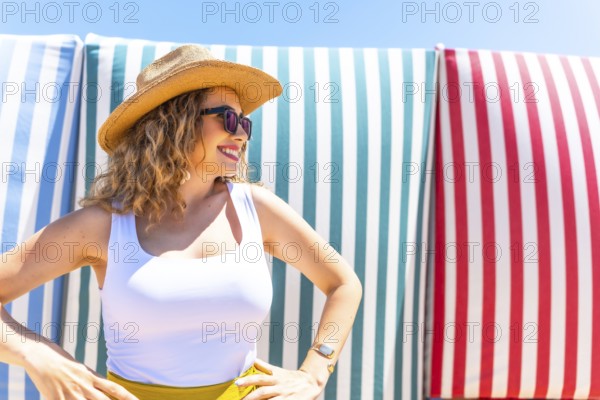 Young woman with curly hair, sunglasses, and straw hat posing with hands on hips in front of colorful beach chairs