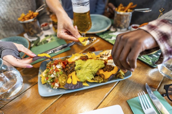 Hands grabbing nachos from plate with guacamole, cheese, jalapenos, and pico de gallo, served on wooden restaurant table