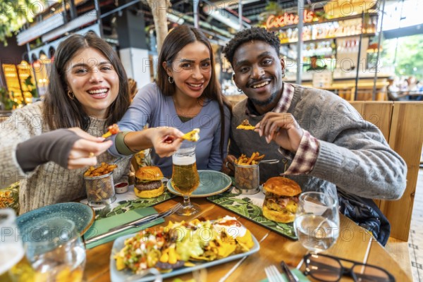Three cheerful multi ethnic friends are enjoying a meal together, sharing nachos, burgers, fries, and beer in a vibrant restaurant setting