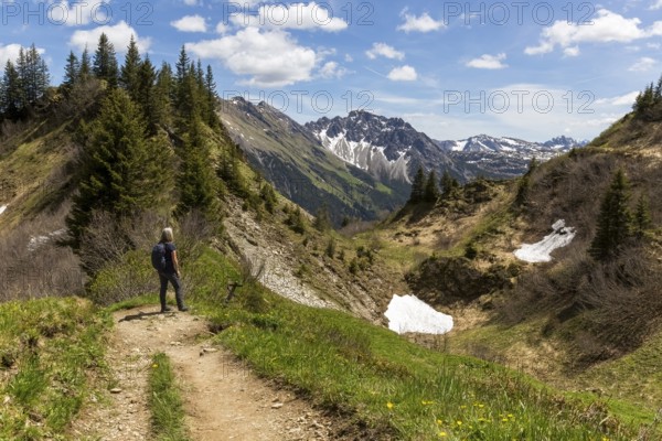 A hiker on a narrow path, surrounded by mountains and scattered snow under a blue sky in Kleinwalsertal, Austria, Europe
