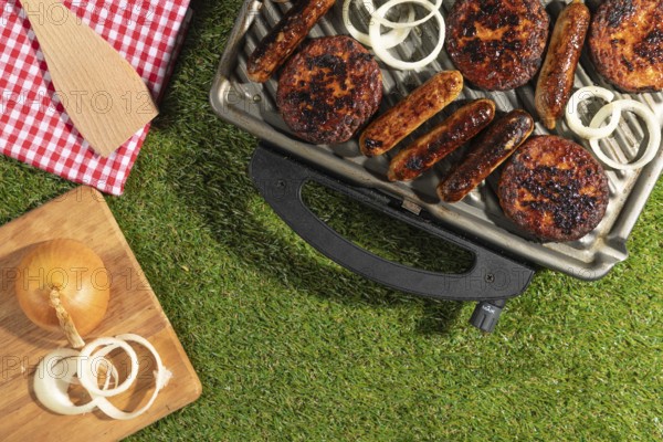 Sausages and burgers on a grill with onions on a wooden board on a green lawn